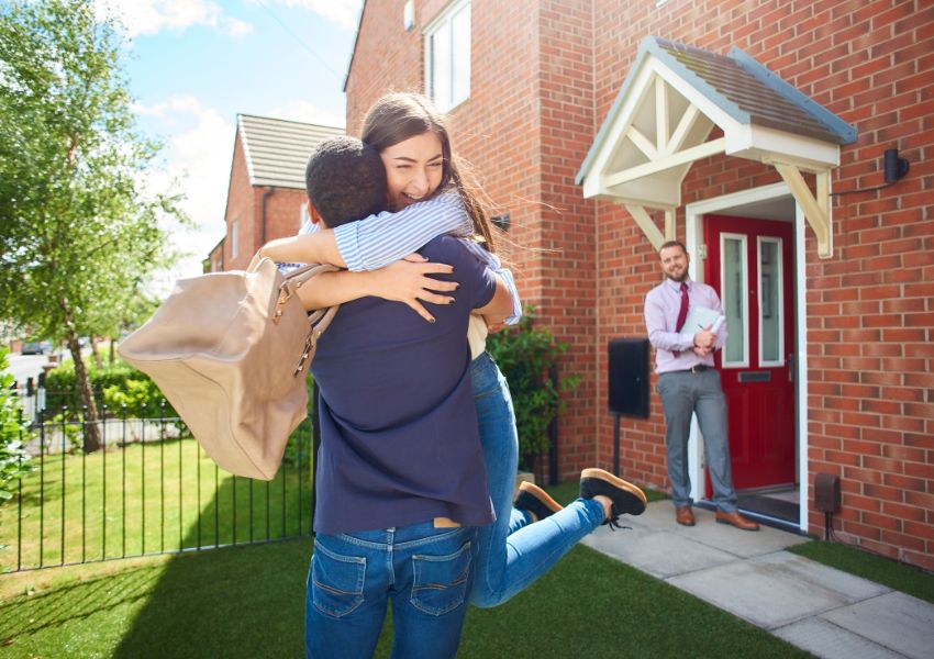two tenant hugging in front of their new rental property as a landlord holds their keys in the background